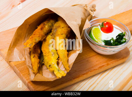 Fish & Chips - Teig - gebratene Sardinen mit klassischen Creme - Käse Sauce und Kräutern auf Holzbrett serviert. Stockfoto