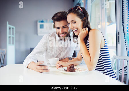 Junge liebende Paar prüfen Telefon zusammen beim Sitzen im Café mit Kaffee und Kuchen Stockfoto
