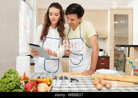 Positive junge hübsche Frau mit Rezept auf Tablet Computer zu Ihrem Freund, wenn Sie zu Hause kochen Stockfoto