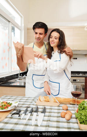 Positive jungen kaukasischen Paar in Schürzen lesen Rezept beim Kochen Buch beim Abendessen zusammen Stockfoto