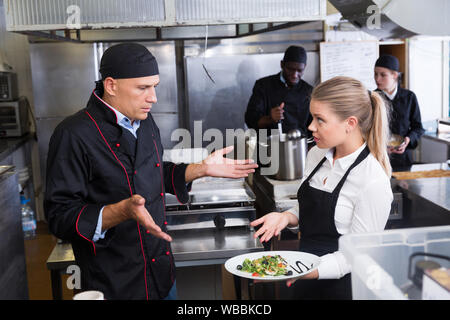 Junge Kellnerin mit gekochte Gericht im Restaurant Küche sprechen mit verwirrten männlichen Chef Stockfoto