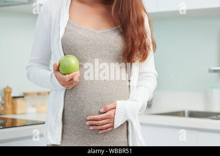 Zugeschnittenes Bild der schwangeren Frau, die ihre grossen Bauch berühren und mit frischem Grün Apple sie hat für Snack Stockfoto