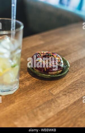 Kalte Limonade mit Eis und Zitronen in ein hohes Glas mit einem Einweg-röhrchen. Schokolade Donut mit Ankleideraum. Lecker und ungesunde Frühstück. Stockfoto