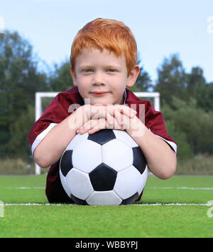 Fröhliche kleine Junge mit Ingwer Haar mit einem Fußball posieren. Porträt eines Kindes in Sportbekleidung. Little Boy liegen auf dem grünen Rasen. Fußball-Weltmeister Stockfoto