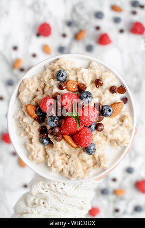 Gesundes Müsli mit Beeren, Schokolade, Mandeln und Honig. Schüssel gehalten in einem womans Hand über einen Marmortisch Hintergrund. Von der Ansicht von oben geschossen Stockfoto