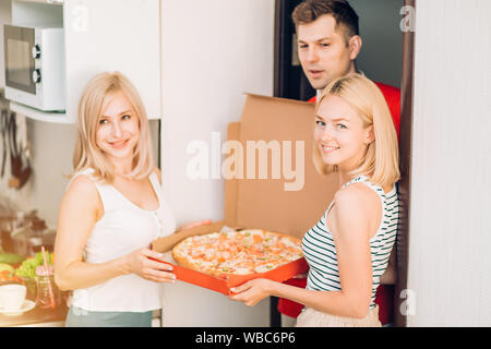 Zwei glückliche Fröhliche kaukasischen weiblichen Studenten die Pizza Delivery man zu Hause. Lieferung Mann in der roten Uniform übersicht Pizza den Kunden am Stockfoto