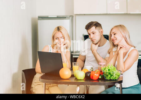 Drei Studenten in den inländischen Kleidung am Tisch sitzen in der Küche, Frühstück vorbereiten, Video Rezept auf Laptop, trinken Orangensaft Stockfoto