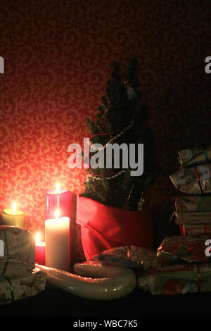 Weihnachtsgeschenke und brennende Kerzen unter einem kleinen Weihnachtsbaum mit roten Hintergrund. Stockfoto