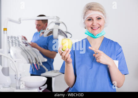 Qualifizierte weibliche stomatologist getrost zu zahnmedizinischen Büro lächelnd, mit frischem Gelb apple Stockfoto