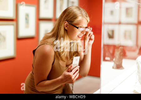 Positive Frau mit Brille und Smartphone Besuch von historischen Museum und Vitrinen mit Exponaten auf der Suche Stockfoto