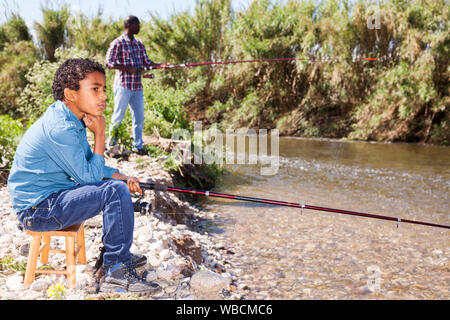 Positive afrikanischer Mann und der kleine Junge in der Nähe von Fluss und Angeln mit Stangen Stockfoto