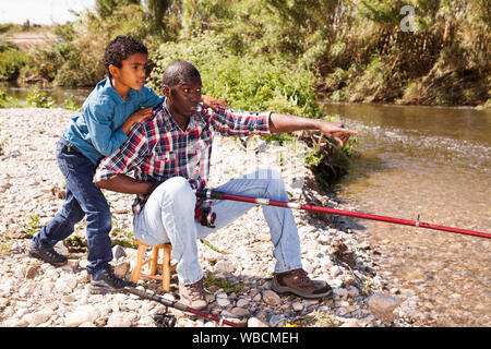 Positive afrikanischer Mann und der kleine Junge in der Nähe von Fluss und Angeln mit Stangen Stockfoto