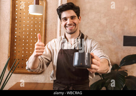 Glückliche junge Mann barista Schürze tragen, der an der Coffee Shop, Kaffeetasse, geben Daumen bis Stockfoto