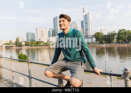 Foto der glückliche junge Mann, Sportswear lächelnd und sitzen auf Geländer, während Sie in der Nähe von City Promenade arbeiten Stockfoto