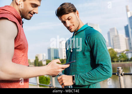 Foto von zwei junge sportliche Männer tragen Sportswear mit Handy und Kopfhörer während der Arbeit aus der Stadt am Flussufer Stockfoto