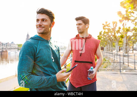 Foto von zwei Stattlichen konzentriert Männer tragen Sportswear mit Handy und Kopfhörer während der Arbeit aus der Stadt am Flussufer Stockfoto