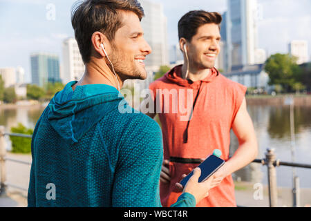 Foto von zwei lächelnde sportliche Männer tragen Sportswear mit Handy und Kopfhörer, während city Riverfront Stockfoto
