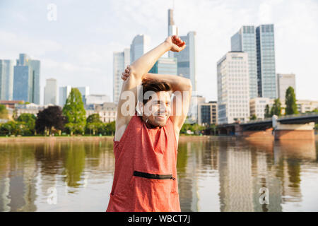 Fotos von lächelnden athletischer Mann mit Sportswear stretching Arm während Sie in der Nähe von City riverfront arbeiten Stockfoto