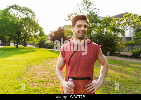 Fotos von lächelnden Mann, sportswear Musik hören mit Kopfhörern und Holding Flasche Wasser, während Sie im City Park arbeiten Stockfoto
