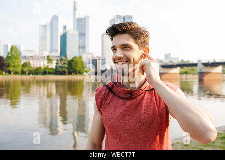 Fotos von lächelnden athletischer Mann mit Sportswear über Kopfhörer, während Sie in der Nähe von City riverfront arbeiten Stockfoto