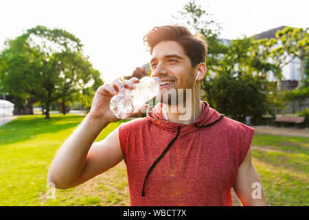 Foto von freudiger Mann, sportswear Musik hören mit Kopfhörern und Trinkwasser während Sie im City Park arbeiten Stockfoto