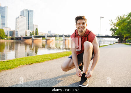 Foto von glücklichen Menschen Sportkleidung tragen mit Ohrhörer und band seine Schnürsenkel, während Sie in der Nähe von City riverfront arbeiten Stockfoto
