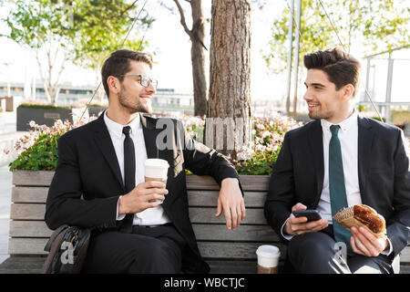 Zwei attraktive lächelnden jungen Unternehmer tragen Anzüge in einer Mittagspause beim Sitzen draußen auf den Straßen der Stadt, Kaffee trinken Stockfoto