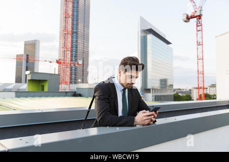 Hübscher junger Unternehmer tragen Anzug wandern draußen auf den Straßen der Stadt und hält Mobiltelefon während lehnte sich auf Schiene Stockfoto