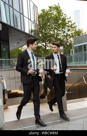 Zwei attraktive selbstbewussten jungen Unternehmer tragen Anzüge wandern draußen auf den Straßen der Stadt, die Tasse Kaffee Stockfoto