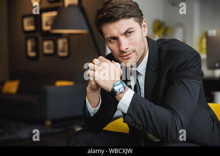 Portrait von selbstbewussten jungen Geschäftsmann tragen formalen schwarzen Anzug und Armbanduhr sitzt auf einem Sessel im Hotel Halle Stockfoto
