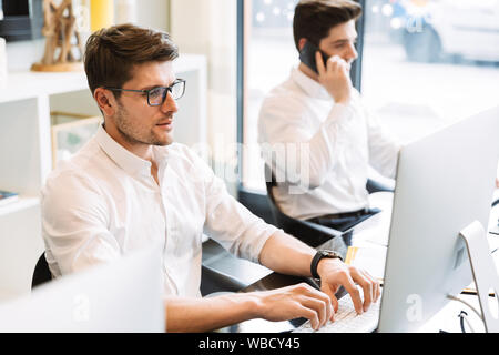 Bild von zwei junge Männer tragen weiße Hemden am Schreibtisch sitzen und Sie Ihr Smartphone während der Arbeit am Computer im Büro Stockfoto