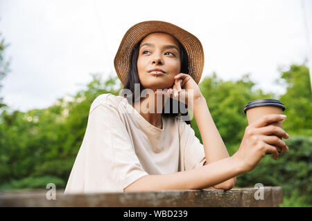 Foto der schönen Frau mit Strohhut und Lippe piercing Holding Pappbecher mit Kaffee zum Mitnehmen im Green Park Stockfoto