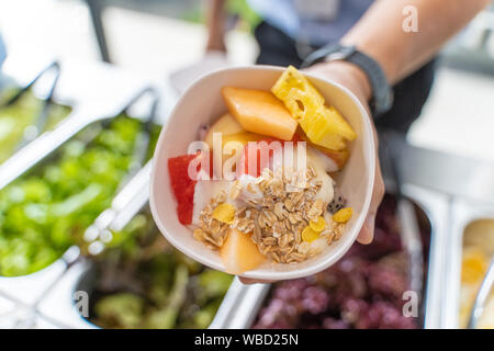 Die Hand des jungen Mann hält eine Schale Obstsalat mit Apfel, Melone, Wassermelone, Ananas, Drachenfrucht, Joghurt und Müsli Stockfoto