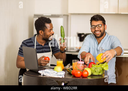 Freudige multi-ethnische Studenten Gesundes Essen und Spaß in der Küche. African American Kerl und sein indischer Freund sitzen am Tisch beobachten Co Stockfoto