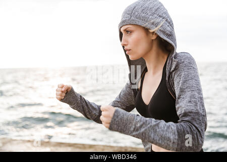 Bild von schweren blonde Frau tragen hoodie Boxen, während die Arbeiten am Pier in der Nähe von Meer in Morgen Stockfoto