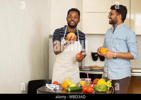 Schuß von zwei männlichen Freunden in Freizeitkleidung in der Küche. Junge Inder und seinem afrikanischen Freund trug Schürze Spaß beim Kochen Abendessen zusammen Stockfoto