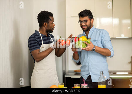 Fröhliche indischen Kerl in Gläser halten Schüssel mit Salat, Paprika und im Gespräch mit den afrikanischen amerikanischen Freund in Schürze Holding reife Tomaten. T Stockfoto