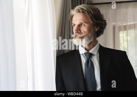 Bild eines bärtigen Reifen gut aussehender Grauhaariger Geschäftsmann posiert im Innenbereich zu Hause in der Nähe der Fenster in formalen gekleidet. Stockfoto