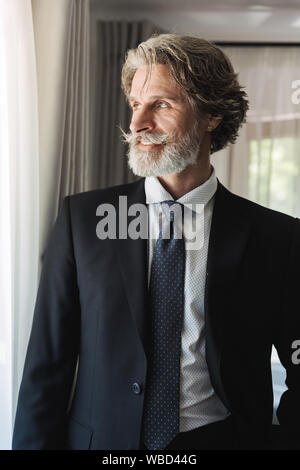 Bild eines bärtigen Reifen gut aussehender Grauhaariger Geschäftsmann posiert im Innenbereich zu Hause in der Nähe der Fenster in formalen gekleidet. Stockfoto