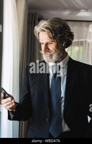 Bild der glücklichen erwachsenen Geschäftsmann das Tragen schwarzer Anzug mit Smartphone und Ohrhörer beim Stehen in der Nähe der Fenster im Hotel Apartment Stockfoto