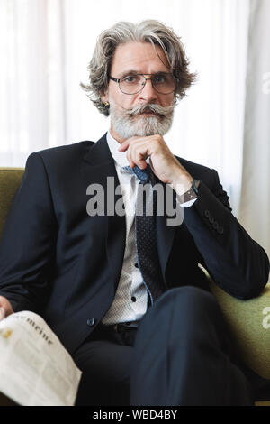 Portrait Nahaufnahme des bärtigen reife Geschäftsmann mit schwarzen Anzug und Brille im Sessel sitzt beim Lesen der Zeitung in der Wohnung Stockfoto
