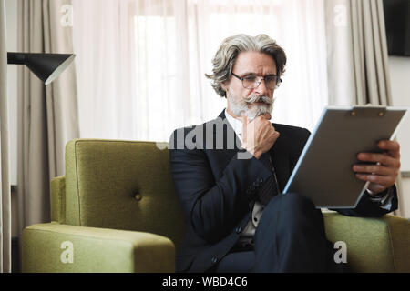 Portrait Nahaufnahme des bärtigen reife Geschäftsmann mit schwarzen Anzug und Brille holding Zwischenablage beim Sitzen auf einem Sessel in der Wohnung Stockfoto