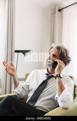 Bild des bärtigen reifer Mann mit Anzug und Krawatte Gespräch am Handy beim Sitzen auf einem Sessel in helles Apartment Stockfoto