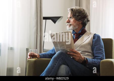 Bild des bärtigen nach Professor Mann mit stilvollen Anzug und Brille lesen Zeitung beim Sitzen auf einem Sessel im Hotel Apartment mit Glas Stockfoto