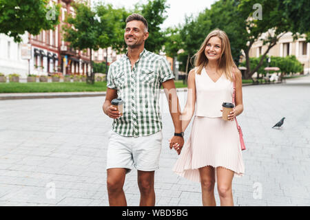 Bild von fröhlichen jungen Paares im Sommer Kleidung und lächelnd die Hände zusammen bei einem Spaziergang durch die Stadt Straße mit Pappbechern Stockfoto