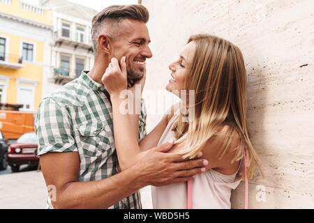 Bild der attraktiven Blondine Paar im Sommer Kleidung lächelnd und zusammen Flirten beim Ständigen gegen die Mauer auf die Stadt. Stockfoto