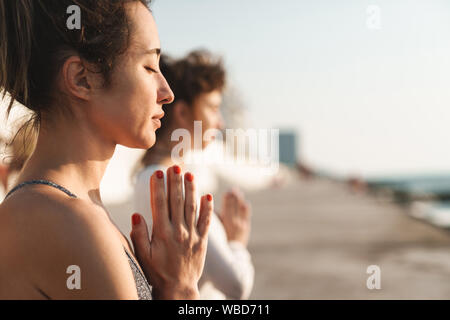 Foto von motiviert junge Frauen in Trainingsanzügen meditieren und die Handflächen zusammen beim Üben von Yoga am Morgen im Freien Stockfoto