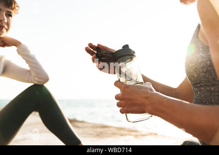 7/8 Image der Jungen fitness Frauen in sportliche Kleidung sitzen auf Yoga Matten mit Wasserflasche von Meer in Morgen Stockfoto