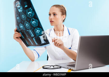 Schönen selbstbewussten jungen Frau Doktor tragen einheitliche an ihrem Arbeitsplatz sitzen, Arbeiten am Laptop über blau Hintergrund isoliert, die Analyse von x Stockfoto