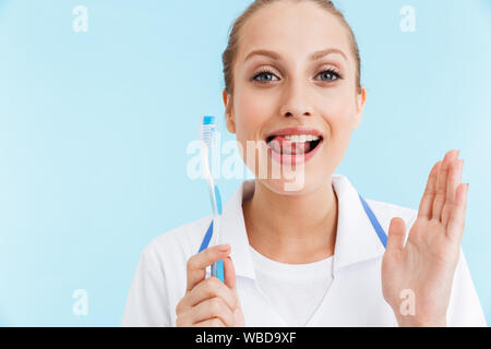 Schöne lächelnde blonde Frau Doktor tragen einheitliche stehend über blauer Hintergrund, weiße teeths und toothsbrush Stockfoto